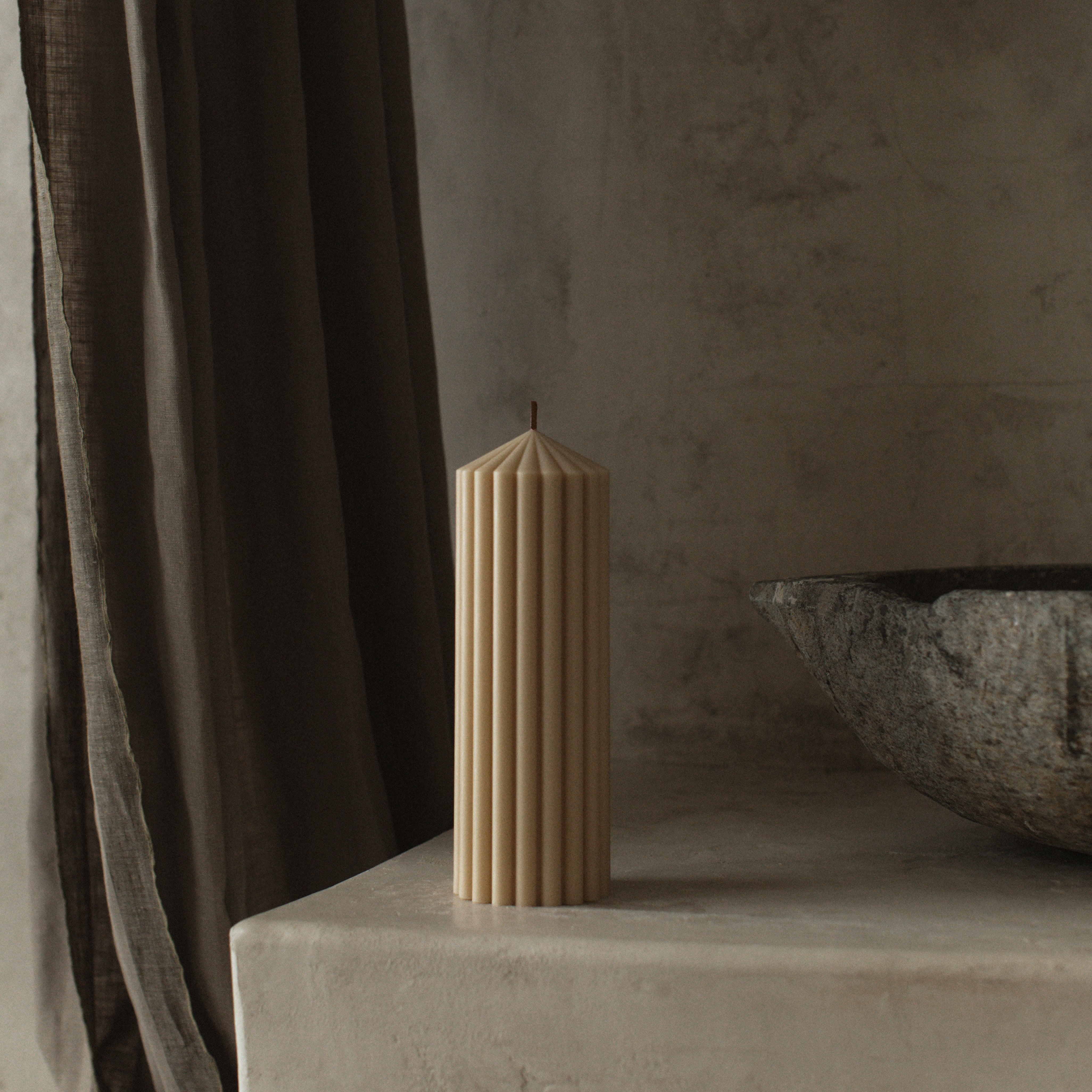 Warm-toned sandstone medium peak candle by LAWA with minimalist mountain peak shape on a concrete surface with a textured wall and curtain in the background