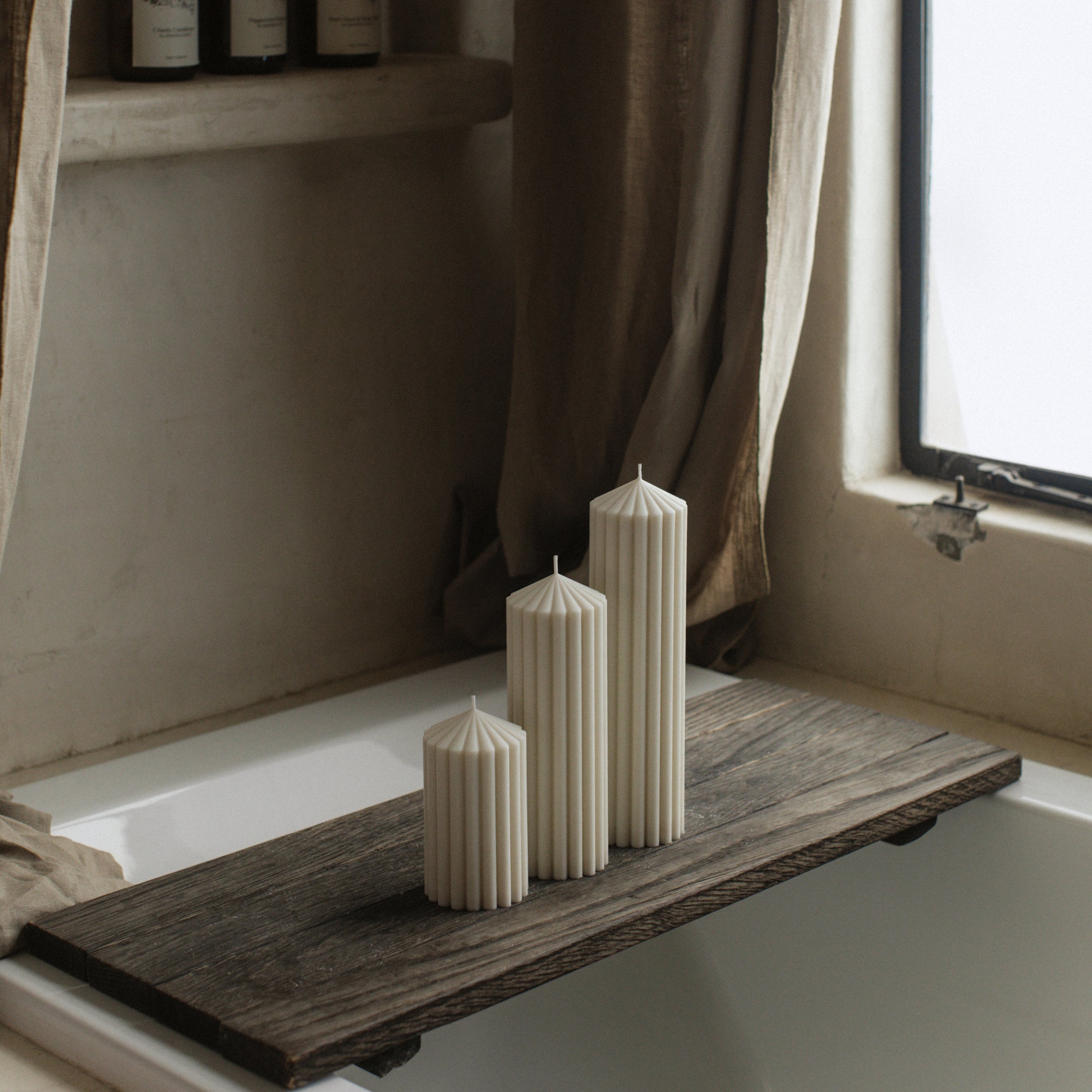LAWA white peak candle trio featuring sculptural mountain-inspired shapes on a wooden tray over a bathtub with a window and curtain in the background.