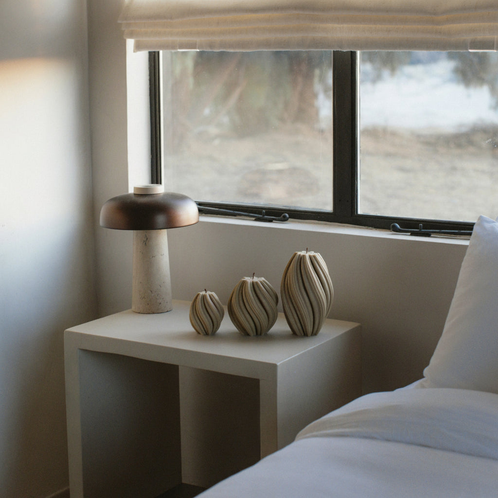 “LAWA swirl pillar candle trio in olive beige featuring sculptural spiral forms in a Modern bedroom with a nightstand featuring a lamp and other decorative items, next to a bed.