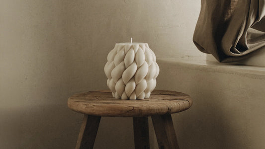 Wide cream sculptural pillar candle with a twisted braided texture on a small rustic wooden stool against a warm neutral wall