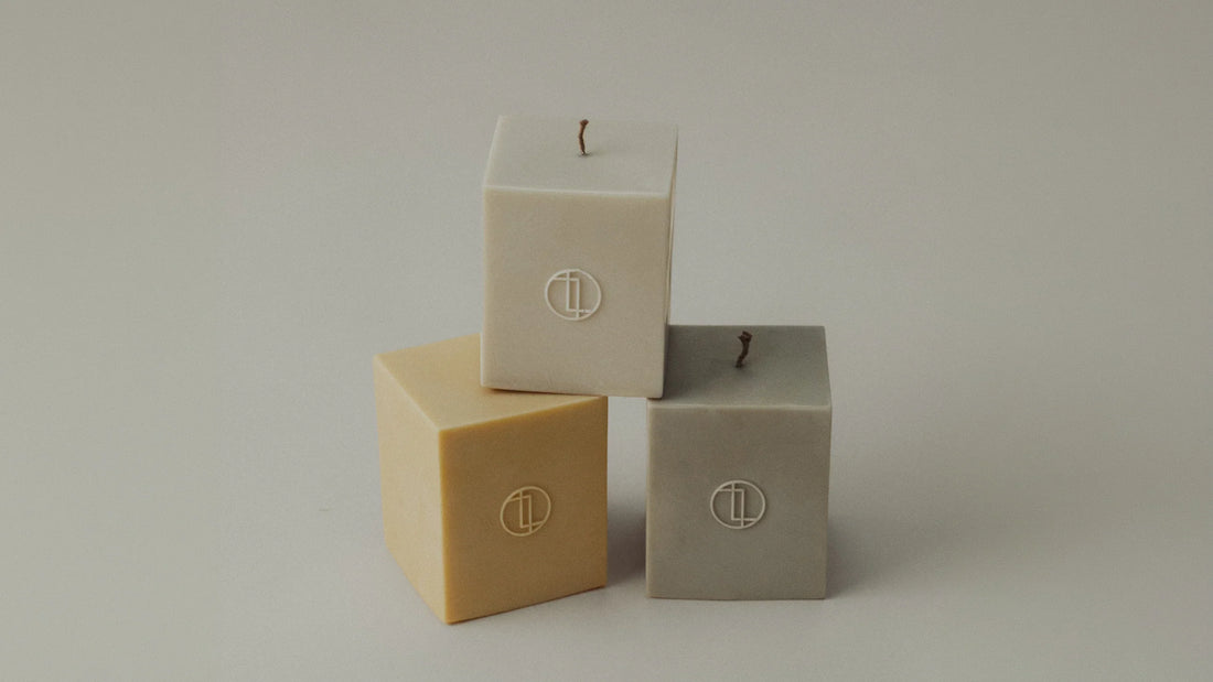 Three cube-shaped pillar candles in cream, butter-beige, and grey, each embossed with the LAWA logo, arranged on a soft neutral background