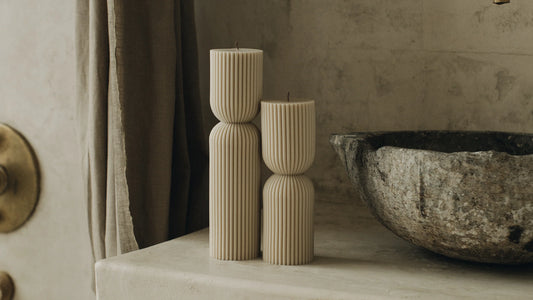Two ribbed beige sculptural pillar candles of different heights on a stone ledge beside a rough stone basin and linen curtain
