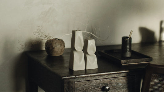 Two cream sculptural pillar candles standing without holders on a dark wooden side table, with a round rattan vase and a black tray in soft natural light