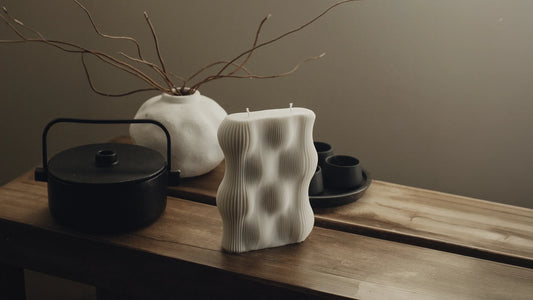 Cream sculptural candle with a wavy, textured surface displayed on a wooden table beside a black teapot, a white branch vase, and a dark tea tray in warm, moody light