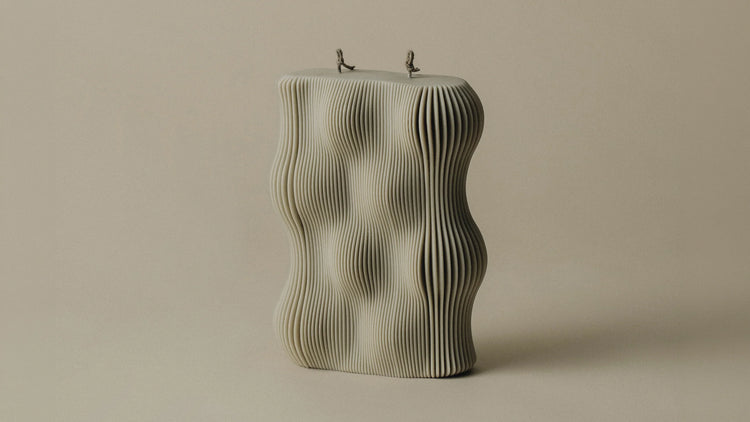 A large wavy pillar candle with two wicks and an undulating rippled texture in sandstone, on a neutral background.