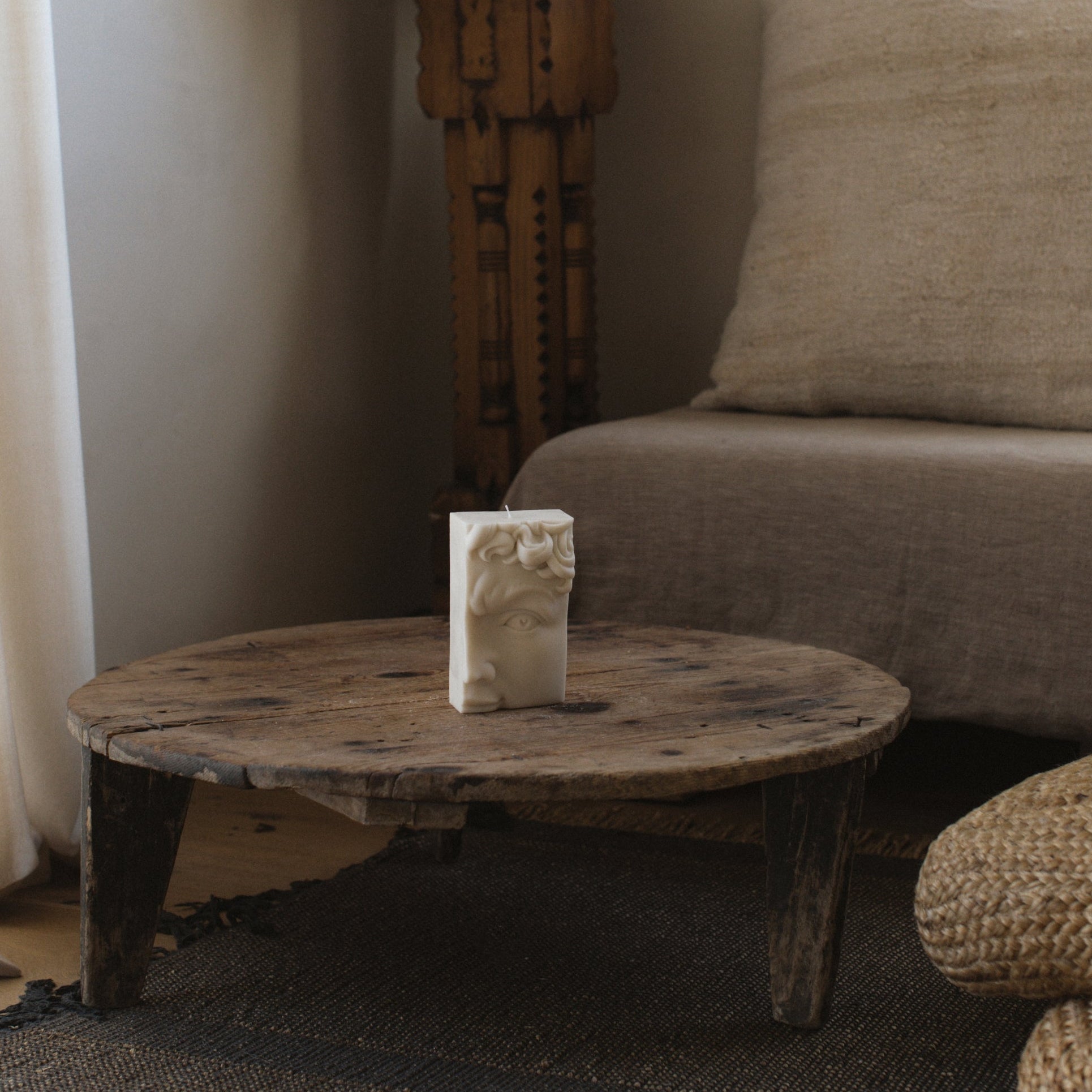 White half-face pillar candle inspired by Michelangelo’s David, sculptural decor by LAWA for a Living room with a wooden table, lamp, and sofa.