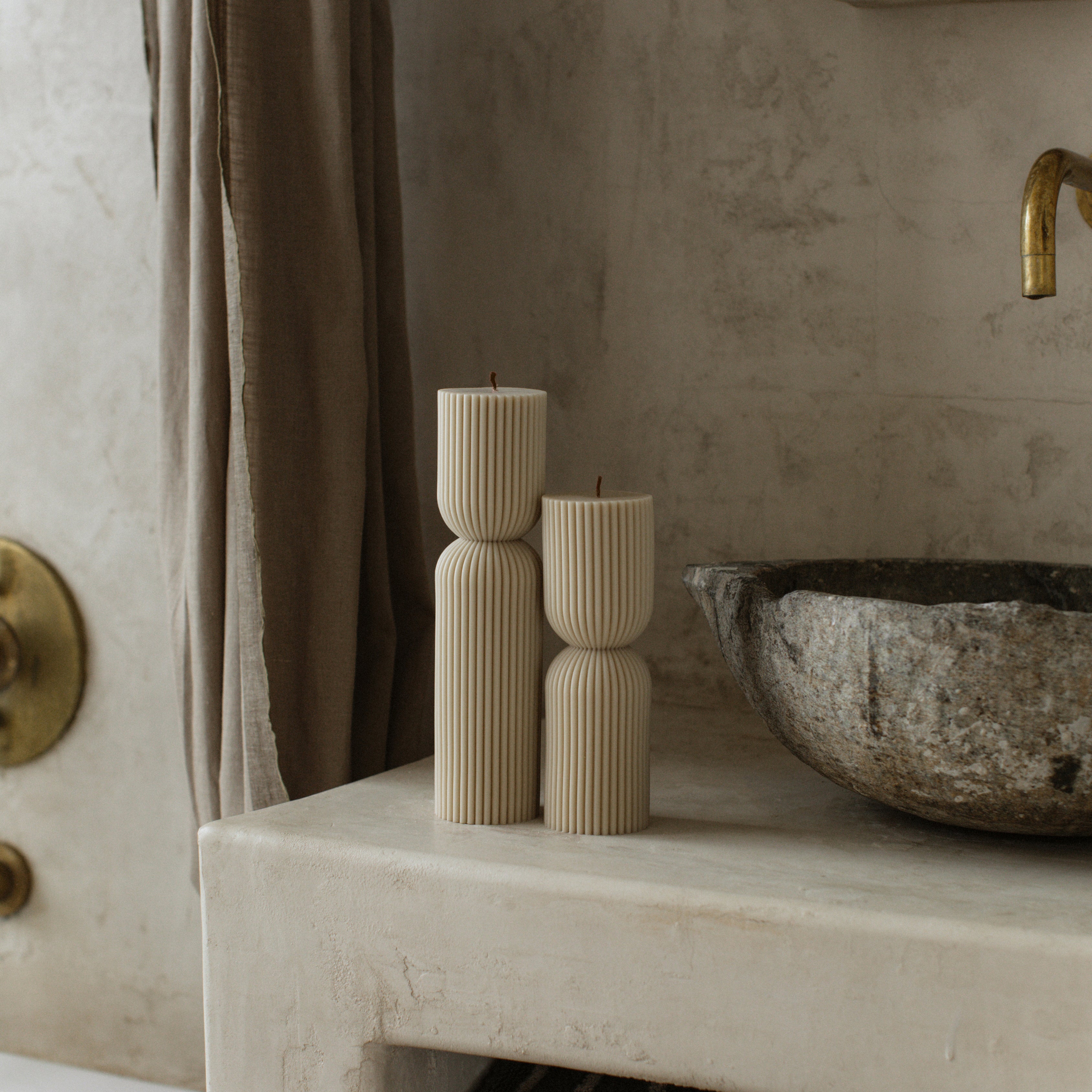 Hourglass set of 2 candles on a stone surface with a textured wall and a curtain in the background.