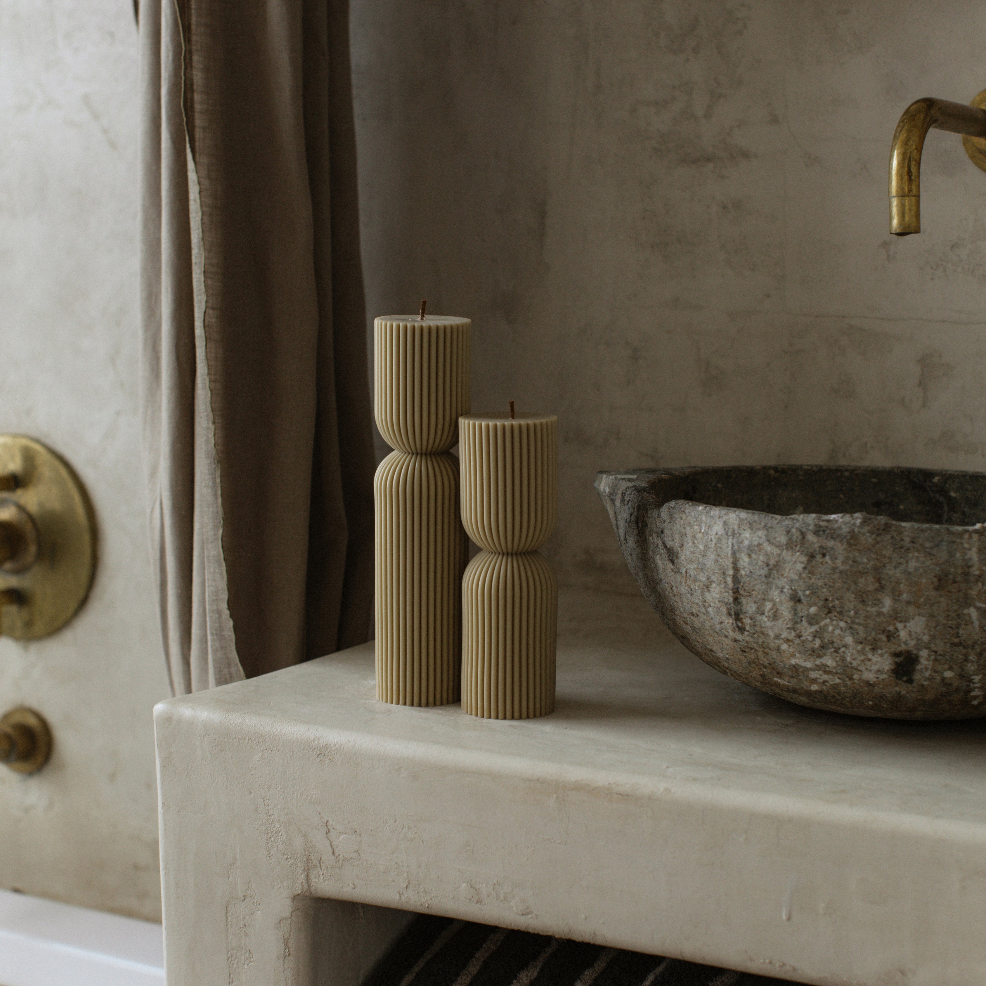 Olive beige hourglass set of two pillar candles  on a stone surface with a textured wall and faucet in the background