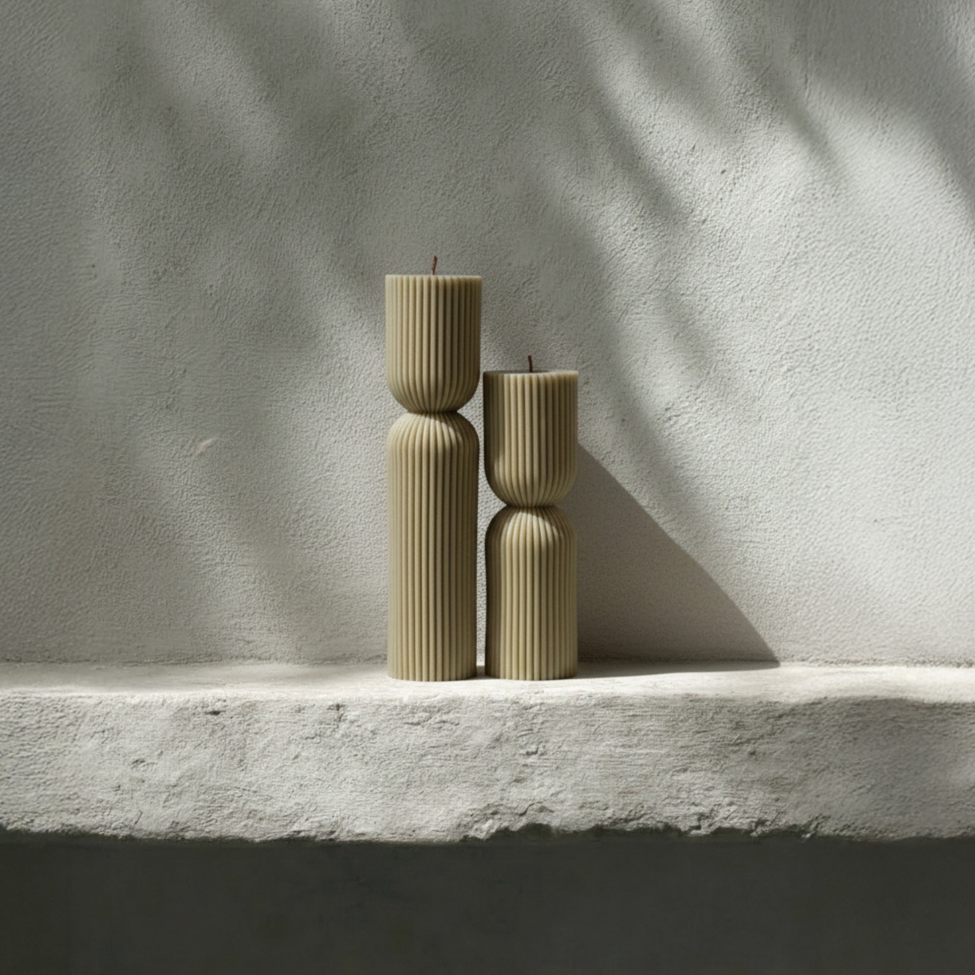 Olive beige hourglass set of two pillar candles on a light stone surface with a soft shadow.