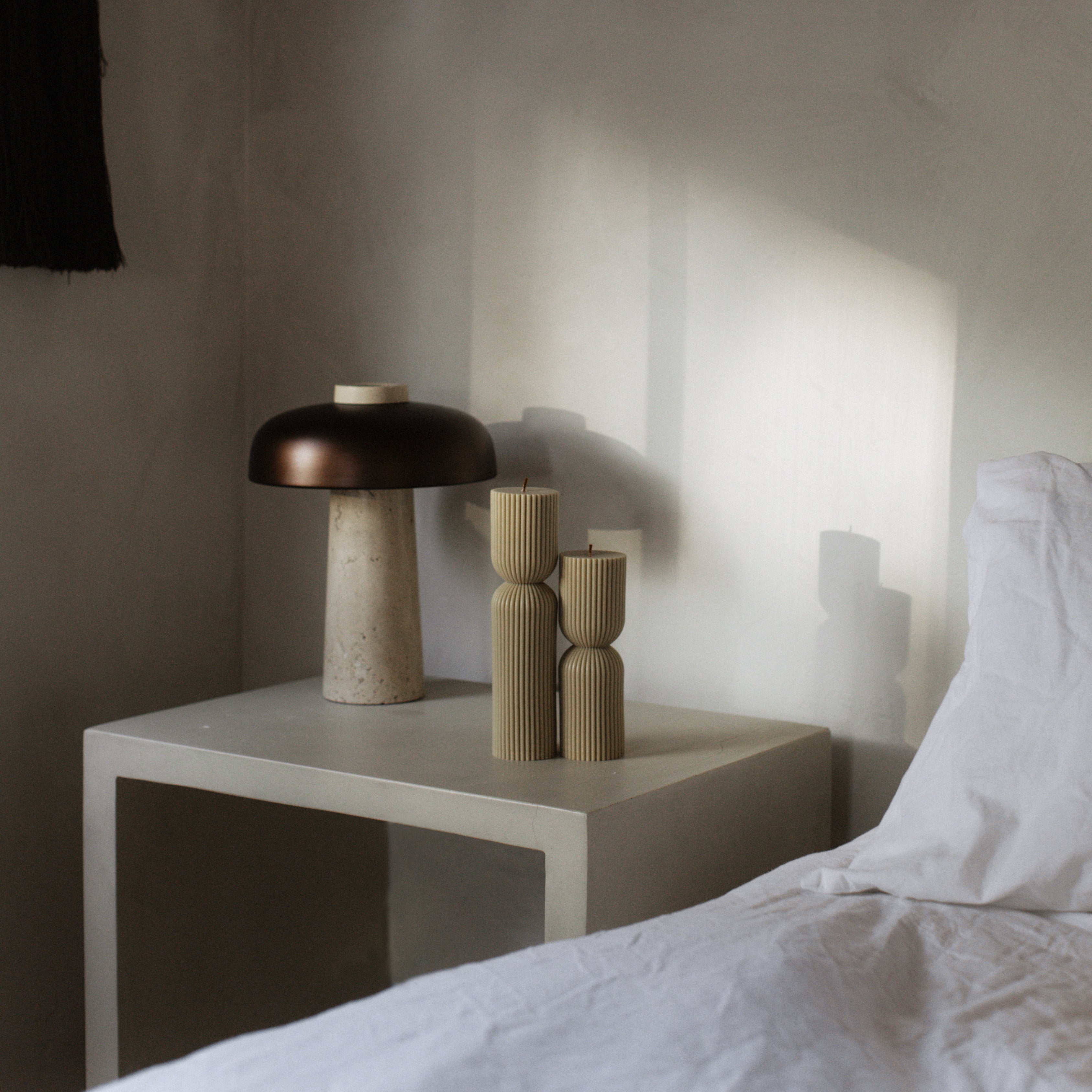Decorative table with a lamp and olive beige hourglass set of two pillar candles next to a bed in a softly lit room.