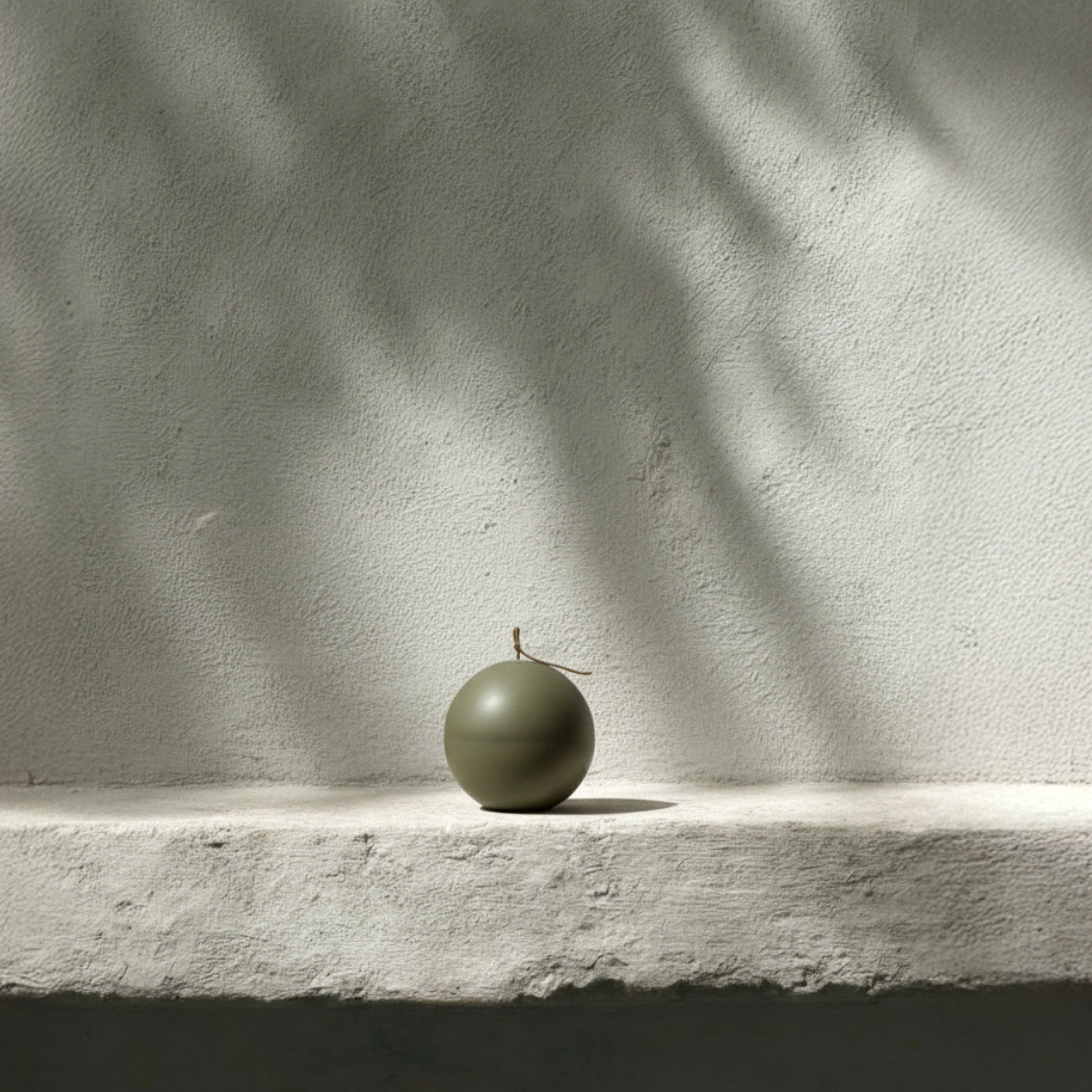 Green imperfect sphere pillar candle on a light surface with shadows cast by leaves on a white wall.