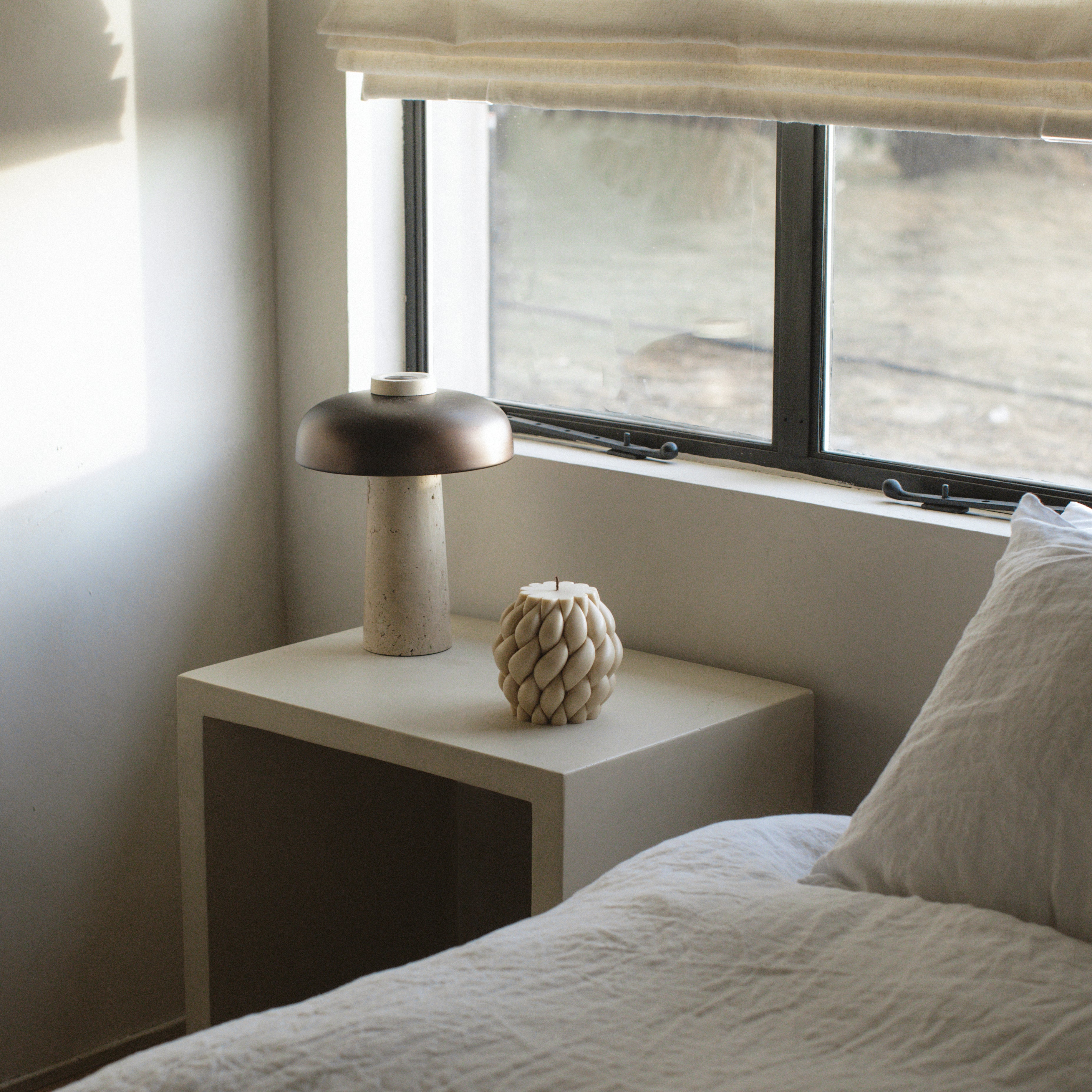 Modern bedroom with a nightstand, lamp, and a moon gray knot ball pillar candle near a window.