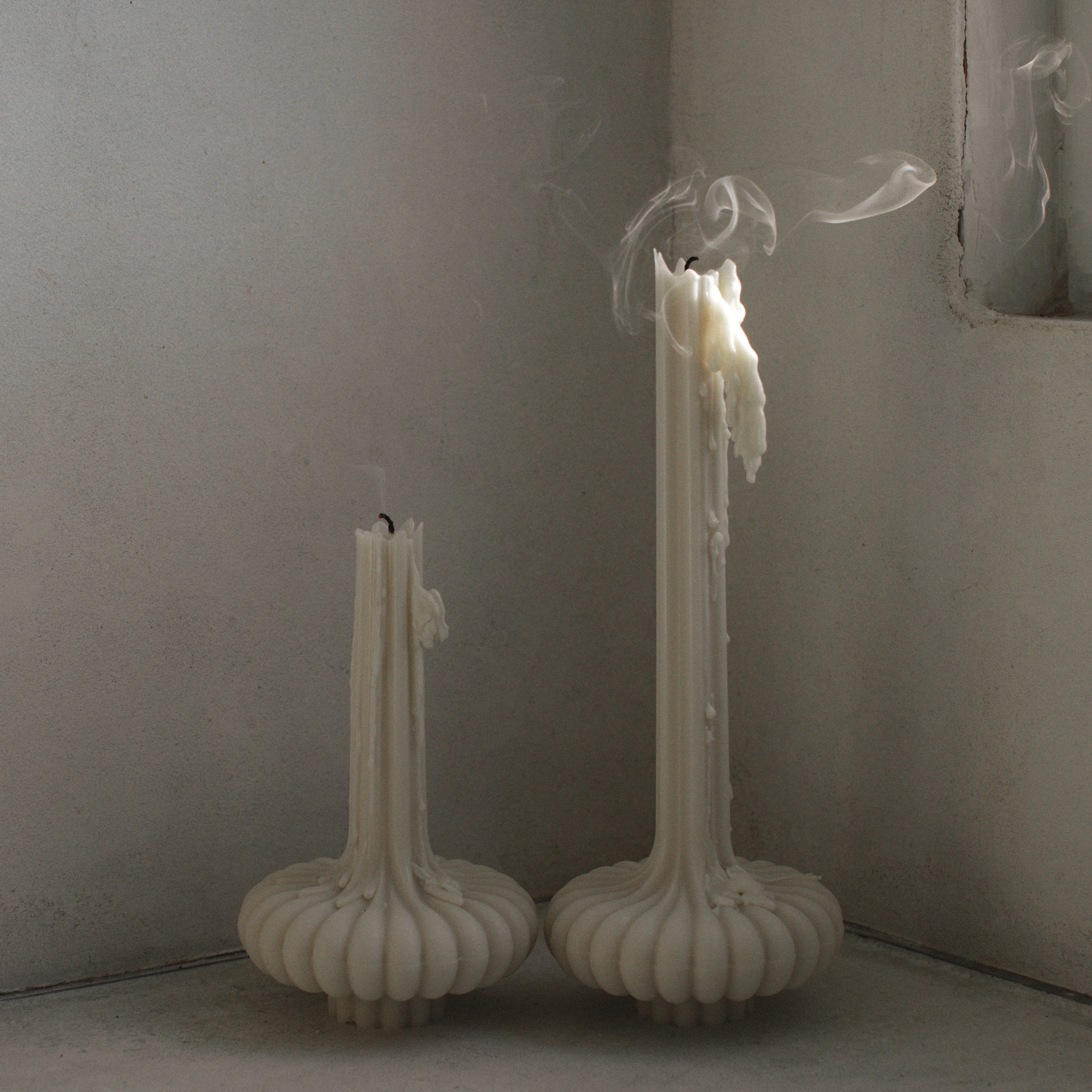 Two white candles with a pumpkin-shaped base, placed on a wooden surface with a textured wall in the background.