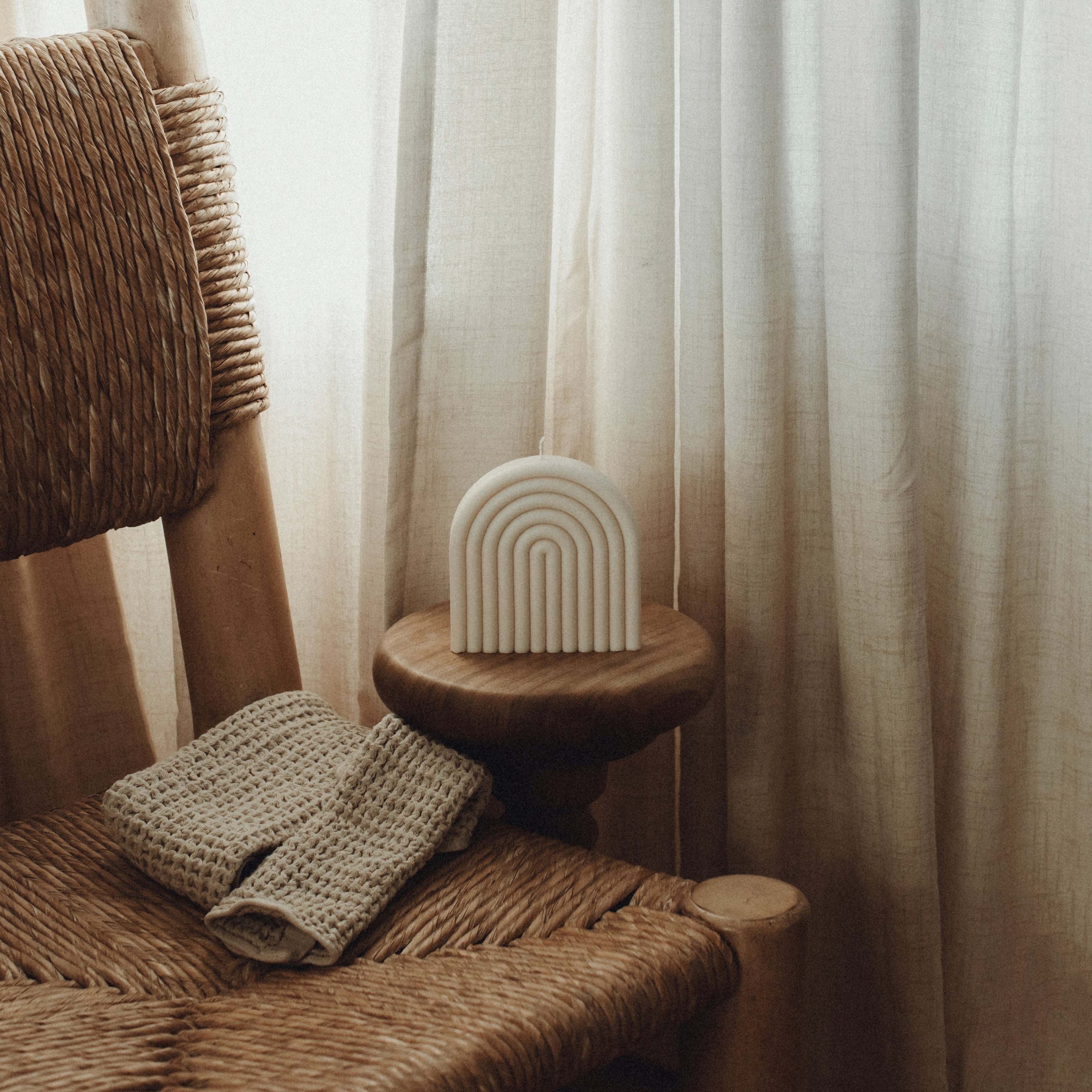 Minimal white rainbow-shaped pillar candle in soy wax by LAWA near a Woven chair with a textured cushion and a small white ceramic arch sculpture against a curtain backdrop