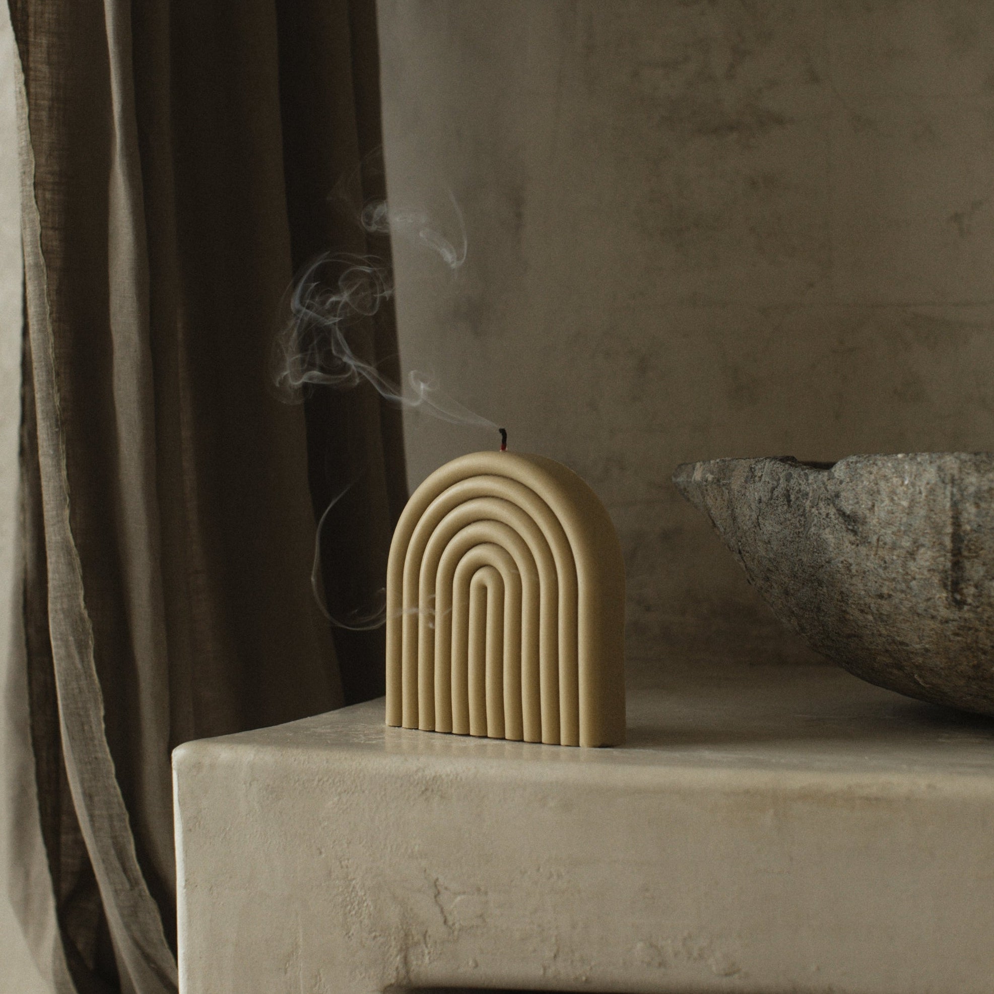 An olive beige, hand-poured soy wax candle in the shape of a rainbow, placed on a concrete surface with smoke rising from its top, indicating it is currently burning.