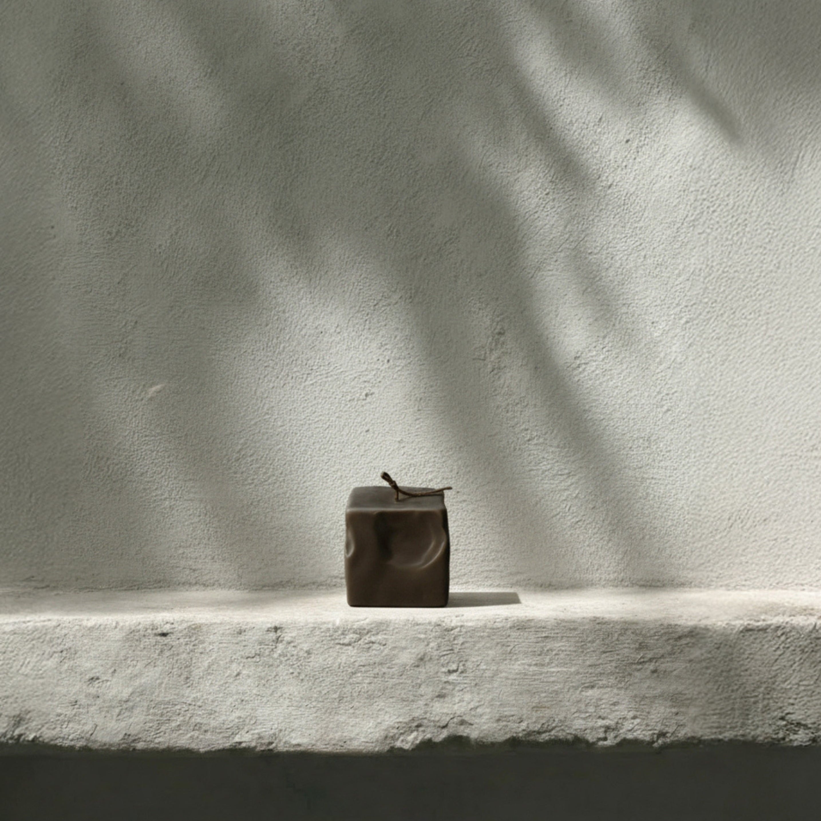 Small kobicha raw block candle by LAWA on a ledge with shadows cast by leaves on a light background