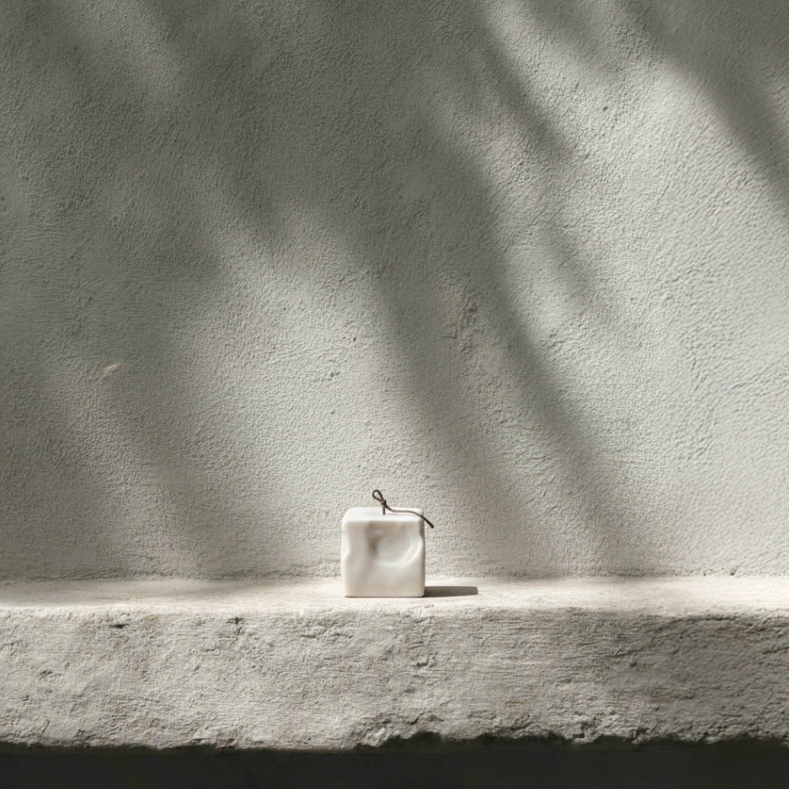 Minimal small light beige raw block pillar candle by LAWA on a concrete ledge with shadows cast by leaves on a light gray wall.