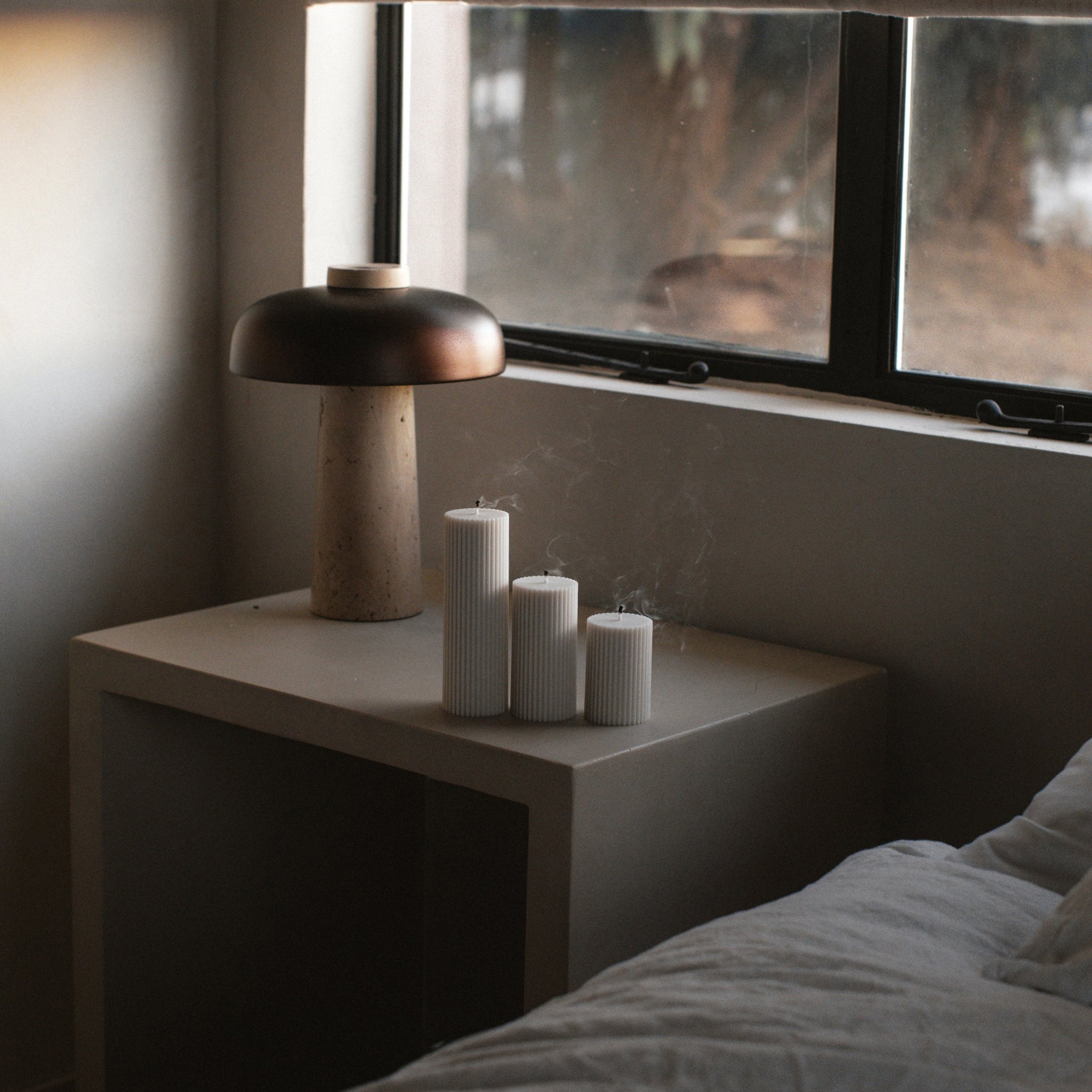 Nightstand with lamp and a set of three 3 ribbed pillar candles near a window in a dimly lit room