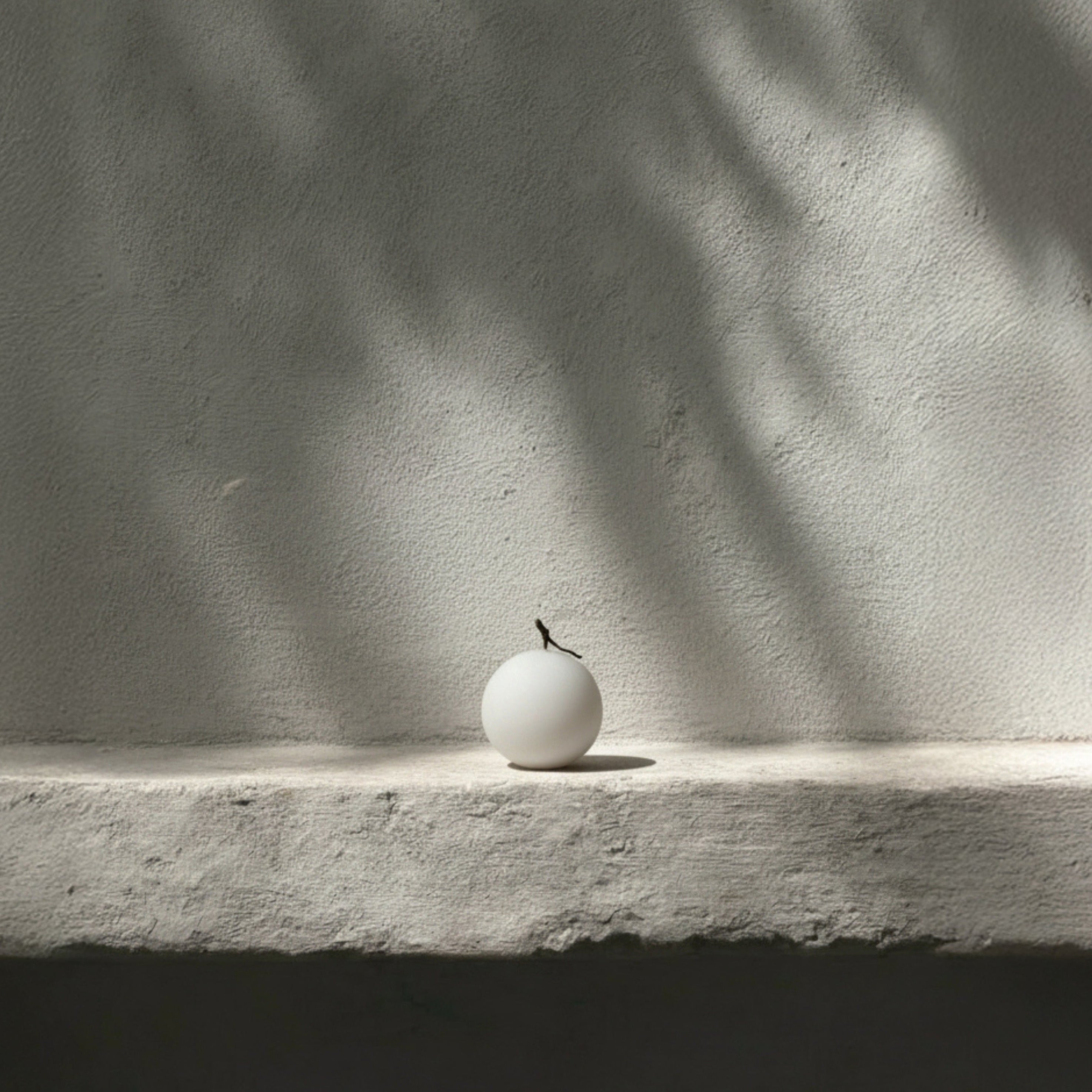 Small white sphere pillar candle on a concrete surface with shadows cast by leaves.
