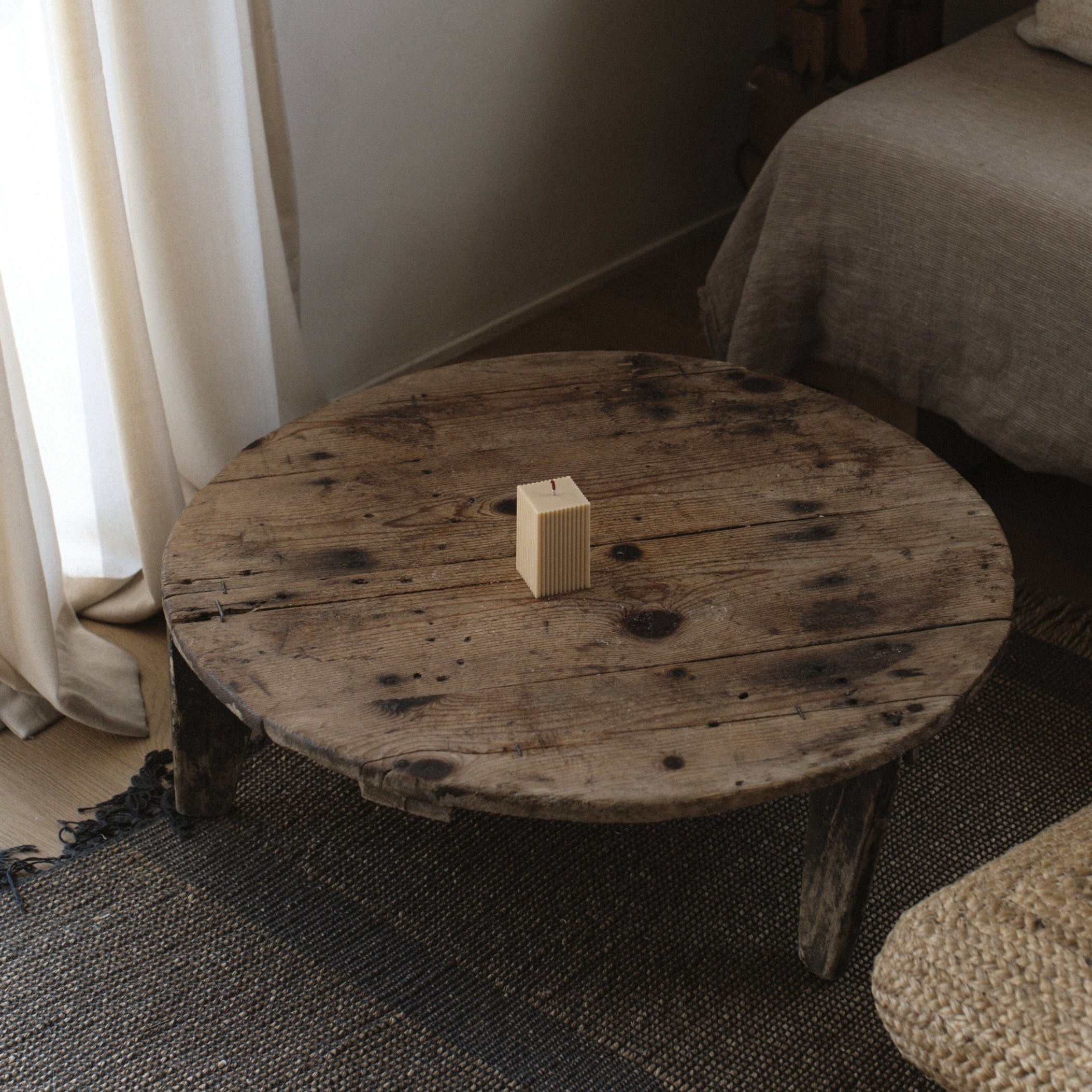 Neutral sandstone ribbed pillar candle by LAWA featuring minimalist grooves on a round wooden table in a room with a curtain and sofa