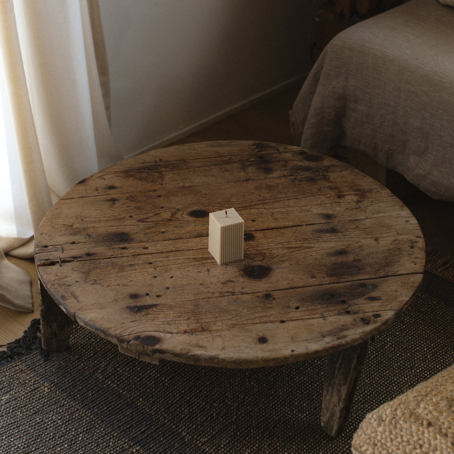 Square ribbed pillar candle in moon gray on a round wooden table in a room with curtains and a sofa.