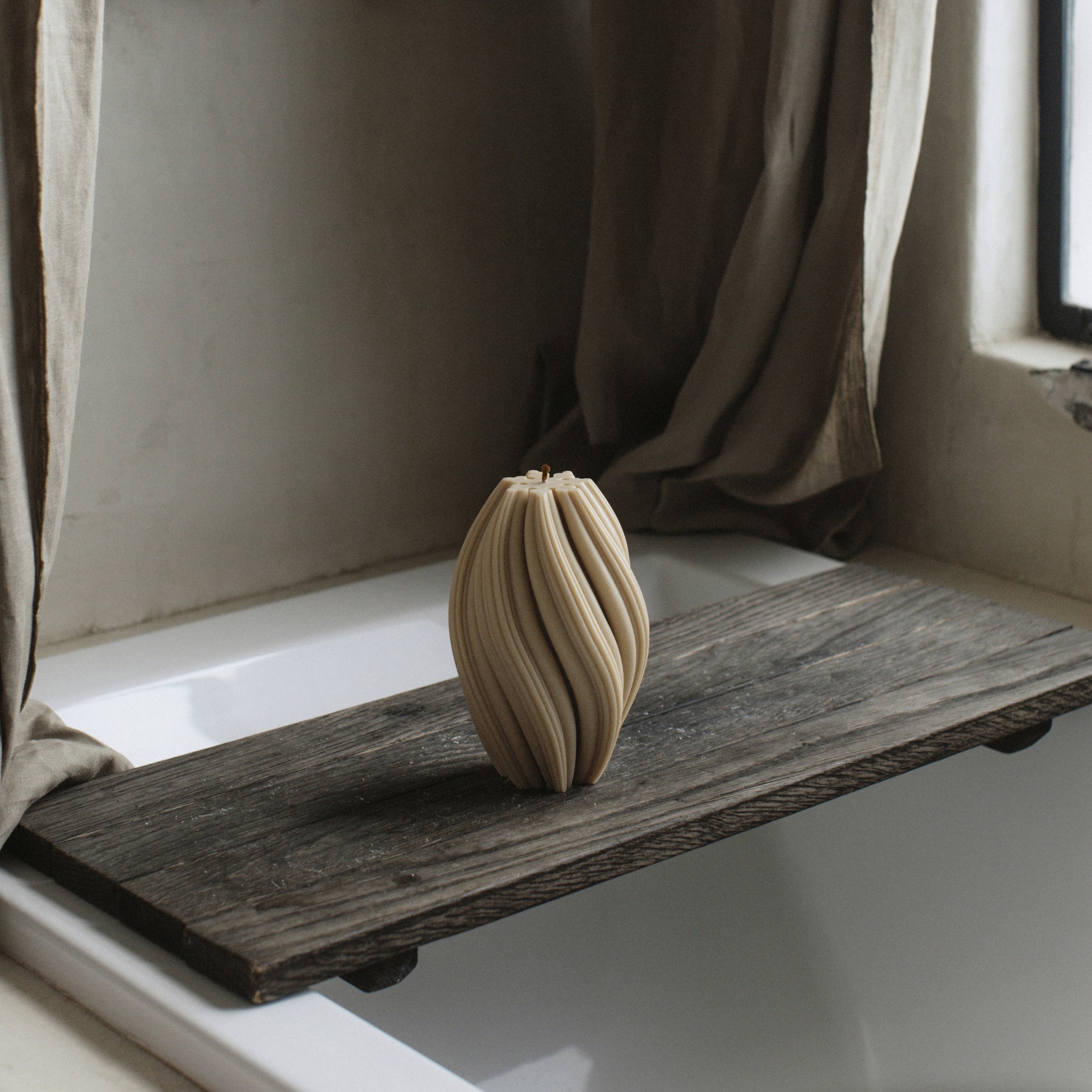 Sandstone large swirl pillar candle by LAWA with elegant curved swirling design on a wooden shelf by a window with curtains.